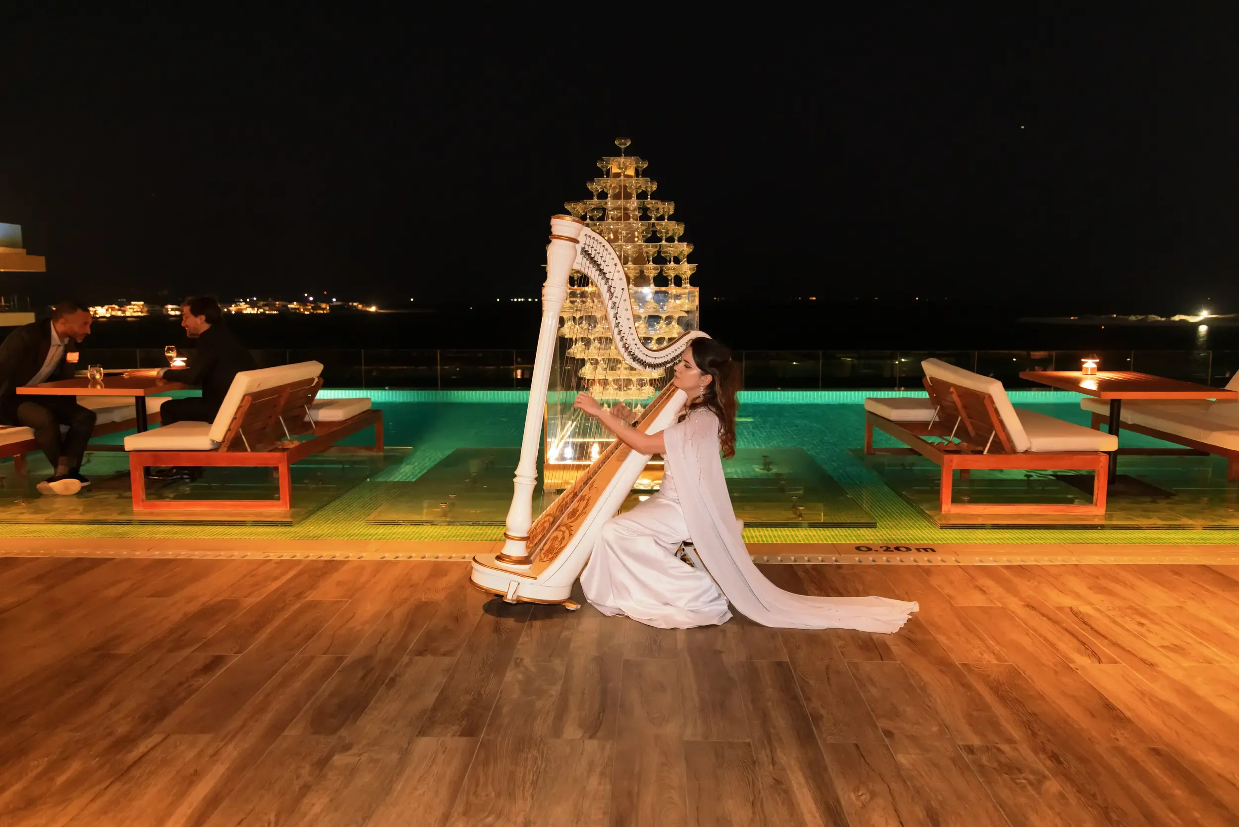 Harpist in a white gown playing beside a champagne tower on a lit pool deck at night