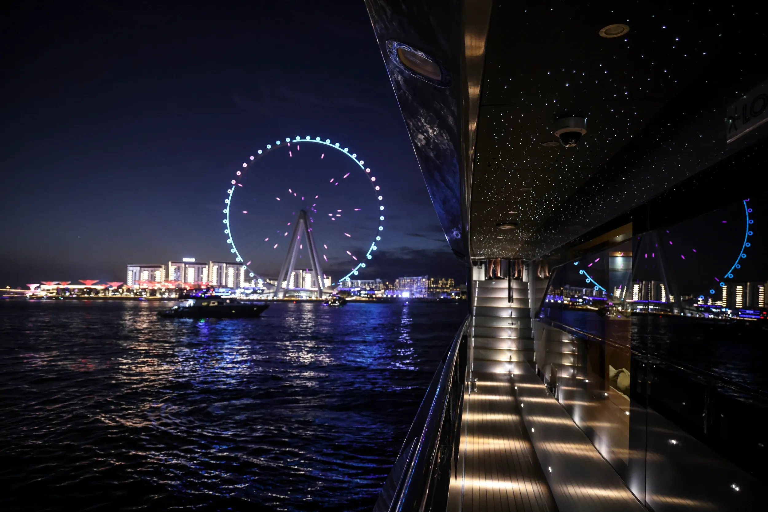 An illuminated yacht walkway beside water with Ain Dubai visible at night.